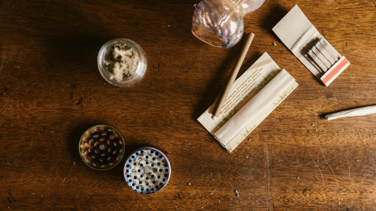 An assortment of cannabis tools (hemp grinder, jar of flower, and rolling paper) on a wooden table